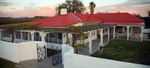 A charming house with a red roof, surrounded by greenery and a white fence, set against a colorful sunset sky.