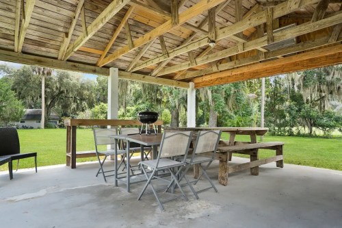 Outdoor pavilion with a wooden roof, picnic tables, and a grill, surrounded by greenery and trees.