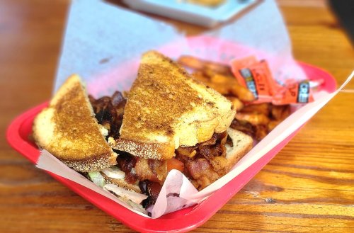 A toasted sandwich with bacon and toppings, served in a red basket with fries and condiment packets.