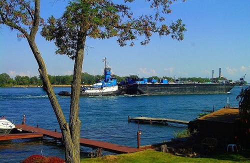 A tugboat navigates a river, pushing a barge past a tree-lined shore and wooden docks under a clear blue sky.