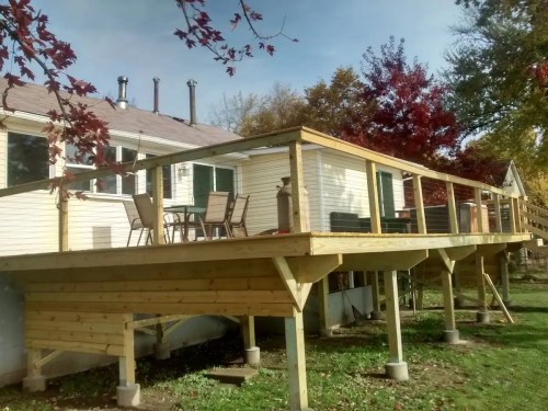 A wooden deck elevated above the ground, attached to a house with chairs and a table, surrounded by trees.