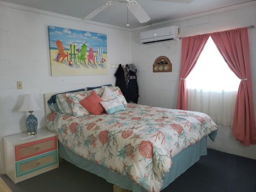 Cozy bedroom with a floral bedspread, a lamp, and beach-themed artwork, featuring a window with light curtains.