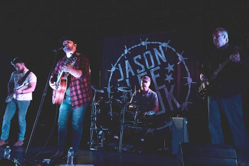 A band performs on stage with a singer holding an acoustic guitar, accompanied by a drummer and two guitarists.