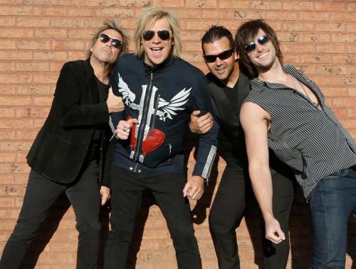 Four musicians pose playfully against a brick wall, wearing sunglasses and stylish outfits, exuding a fun vibe.