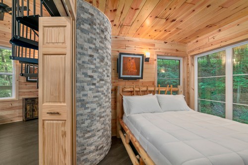 Cozy wooden bedroom with a bed, stone accent wall, and spiral staircase, surrounded by large windows and greenery.