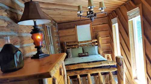 Cozy log cabin interior featuring a wooden bed, lamp, and large windows with natural light.