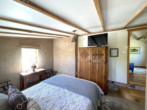 Cozy bedroom with a wooden bed, decorative quilt, and a TV, featuring rustic walls and natural light from a window.