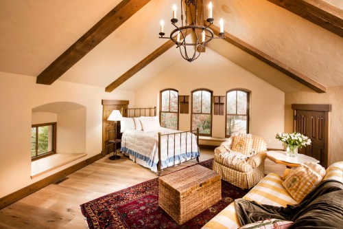 Cozy bedroom with wooden beams, a bed, armchair, and a decorative rug, featuring large windows and natural light.