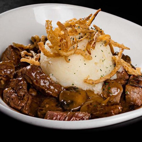 A bowl of beef tips in gravy served over mashed potatoes, topped with crispy fried onions.