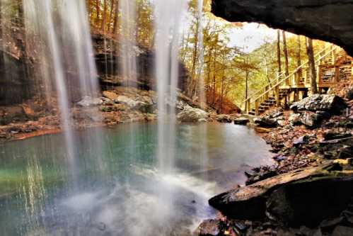 A serene waterfall cascades into a clear pool, surrounded by autumn foliage and rocky terrain. A wooden path is visible nearby.