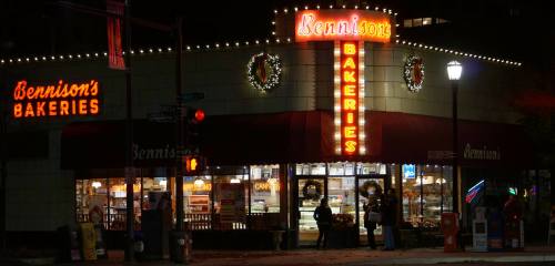 A brightly lit bakery at night, featuring festive wreaths and a sign that reads "Bennison's Bakeries."
