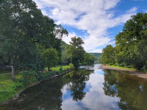 A serene river scene surrounded by lush trees and a blue sky with fluffy clouds reflecting on the water's surface.