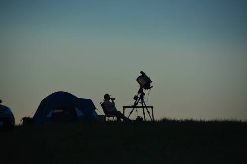 A silhouette of a person sitting in a chair next to a telescope and tent against a twilight sky.