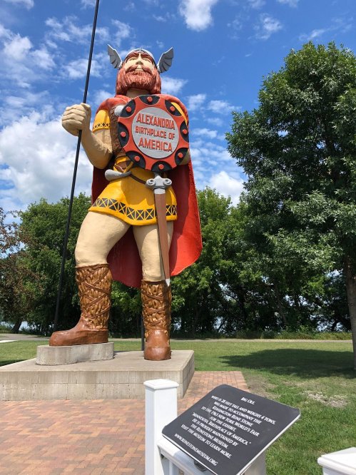 A large statue of a Viking warrior stands proudly, holding a spear, with a plaque nearby in a park setting.