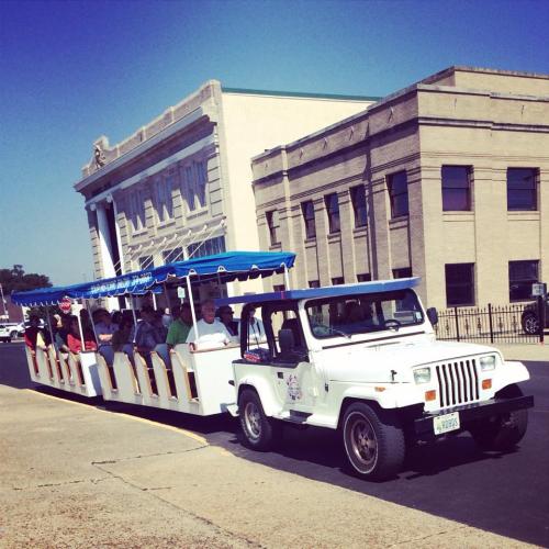 A white tram with passengers rides along a street near a historic building under a clear blue sky.