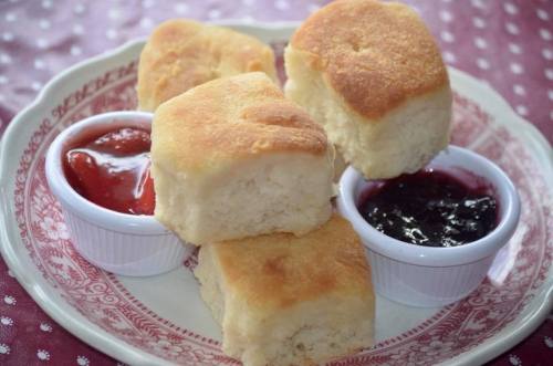 A plate of golden-brown biscuits served with small bowls of strawberry and grape jam.