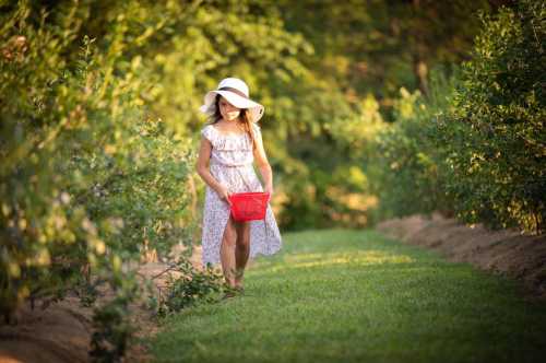 A young girl in a white dress and hat walks through a green field, carrying a red basket.