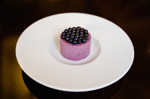 A round purple dessert topped with glossy black berries, served on a white plate against a dark background.