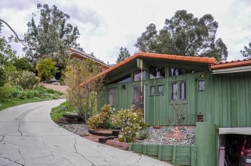 A green house with a red tile roof, surrounded by plants, on a winding driveway with cloudy skies above.