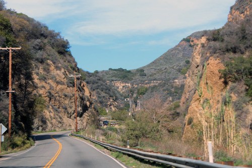 A winding road through a hilly landscape with greenery and rocky cliffs under a clear blue sky.