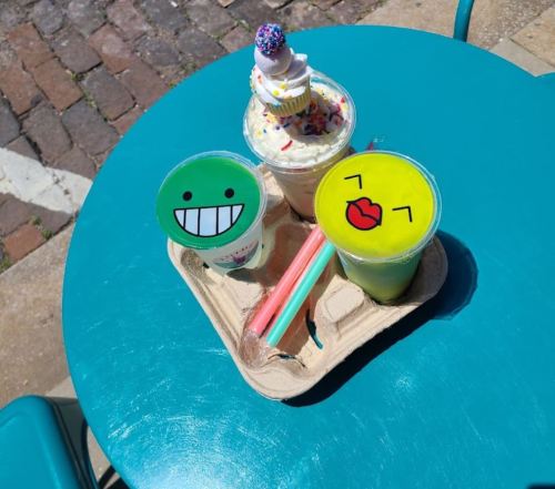 Three colorful drinks with playful smiley faces on the cups, served on a tray with two straws, on a teal table.