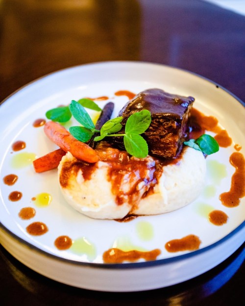 A plate of tender beef with rich sauce, served on creamy mashed potatoes and garnished with carrots and fresh herbs.