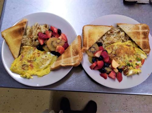 Two plates of breakfast: one with an omelet, toast, hash browns, and fruit; the other with a colorful omelet and similar sides.