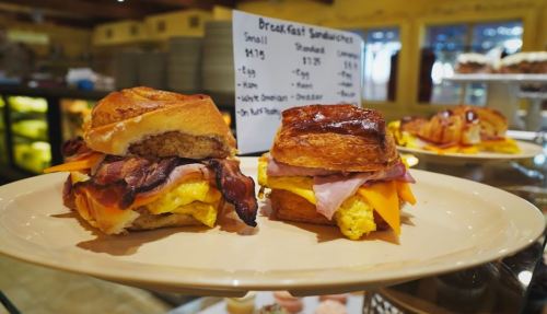 Two breakfast sandwiches on a plate, featuring eggs, cheese, ham, and bacon on toasted buns, with a menu in the background.