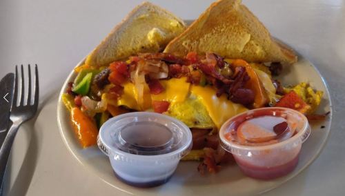 A plate of omelette topped with cheese, bacon, and vegetables, served with toast and two dipping sauces.