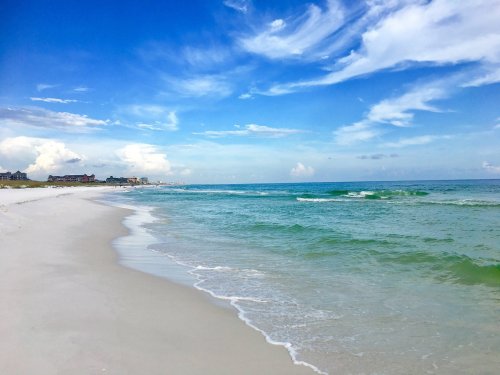 A serene beach scene with soft white sand, gentle waves, and a bright blue sky with scattered clouds.
