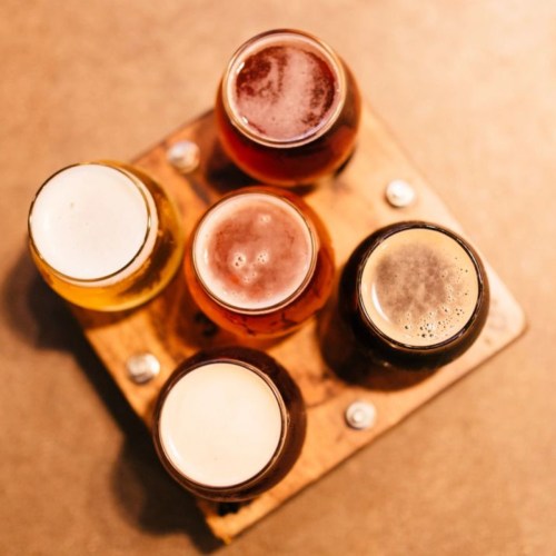 A wooden flight board with five different glasses of beer, showcasing various colors and foam levels.