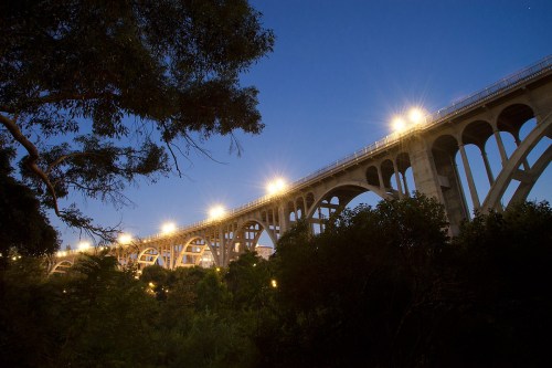 A lit bridge arches over a lush landscape at dusk, framed by trees and a clear blue sky.
