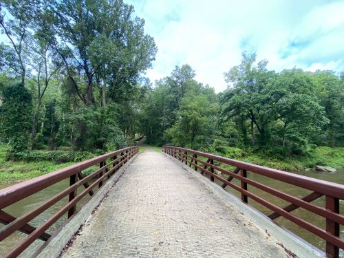 A wooden bridge spans a calm river, surrounded by lush green trees and a cloudy sky.