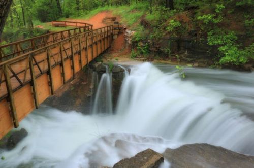 A wooden bridge spans a flowing waterfall surrounded by lush green trees and rocky terrain.