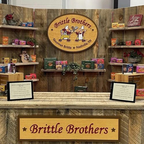 A wooden display featuring "Brittle Brothers" gourmet brittle products, with festive decorations and signage.