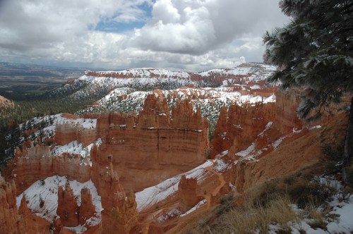 A scenic view of red rock formations and snow-capped peaks under a cloudy sky in a mountainous landscape.