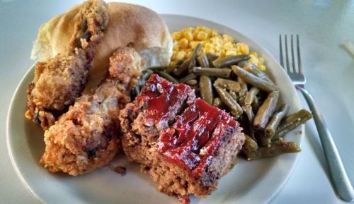 A plate of fried chicken, meatloaf with glaze, green beans, corn, and a roll.