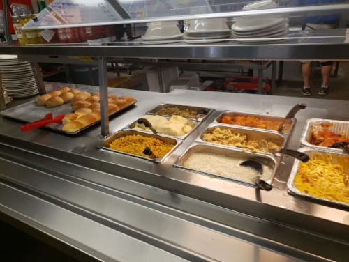 A cafeteria food line featuring rolls, macaroni and cheese, rice, pasta, and orange chicken in metal trays.