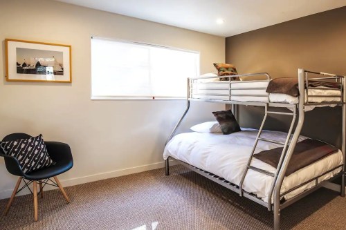A cozy bedroom featuring a bunk bed, a single bed, and a chair, with natural light from a window.