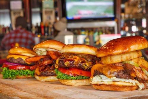 Four gourmet burgers stacked on a wooden board, featuring fresh ingredients and toppings, with a bar scene in the background.