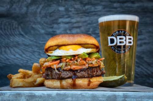 A juicy burger topped with an egg and veggies, served with fries and a pint of beer in the background.