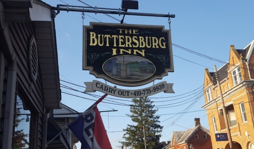 Sign for The Buttersburg Inn, featuring "Carry Out" and contact number, with buildings and trees in the background.