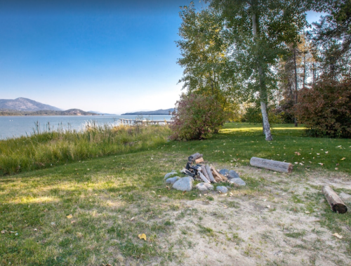 A serene lakeside scene with a fire pit, grassy area, and trees under a clear blue sky. Mountains are visible in the distance.