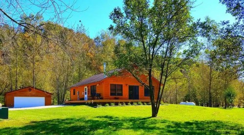 A cozy orange cabin surrounded by trees and a green lawn on a sunny day. A small shed is nearby.