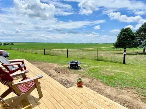 A scenic view from a wooden deck overlooking green fields and a blue sky with fluffy clouds. A fire pit is visible.