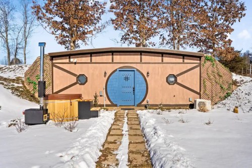 A cozy, round building with a blue door, surrounded by snow and trees, featuring a stone pathway and outdoor hot tub.