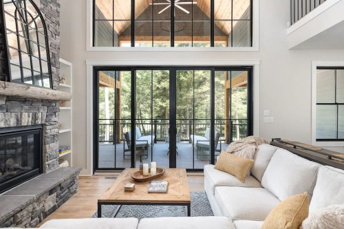 Modern living room with a stone fireplace, large windows, and a view of a deck surrounded by trees. Cozy and inviting atmosphere.
