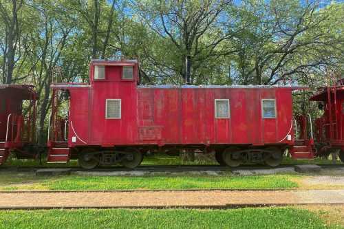 A red caboose parked on a track, surrounded by green trees and grass under a clear blue sky.