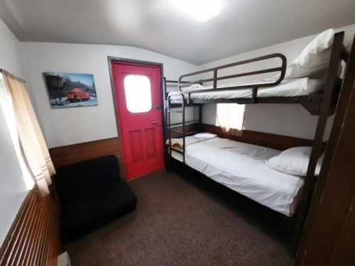 A small room with a bunk bed, single bed, and a red door, featuring a cozy seating area and a wall painting.