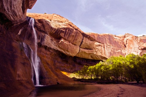 A serene waterfall cascades down rocky cliffs, surrounded by lush greenery and a sandy riverbank under a clear sky.